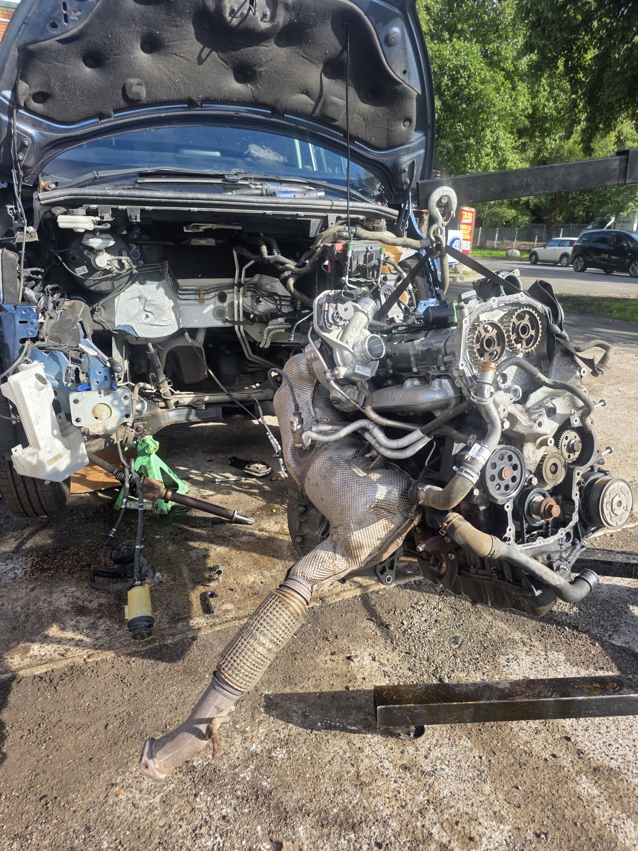 An engine lifted out of a car on a driveway, with the cylinder head and timing gears exposed