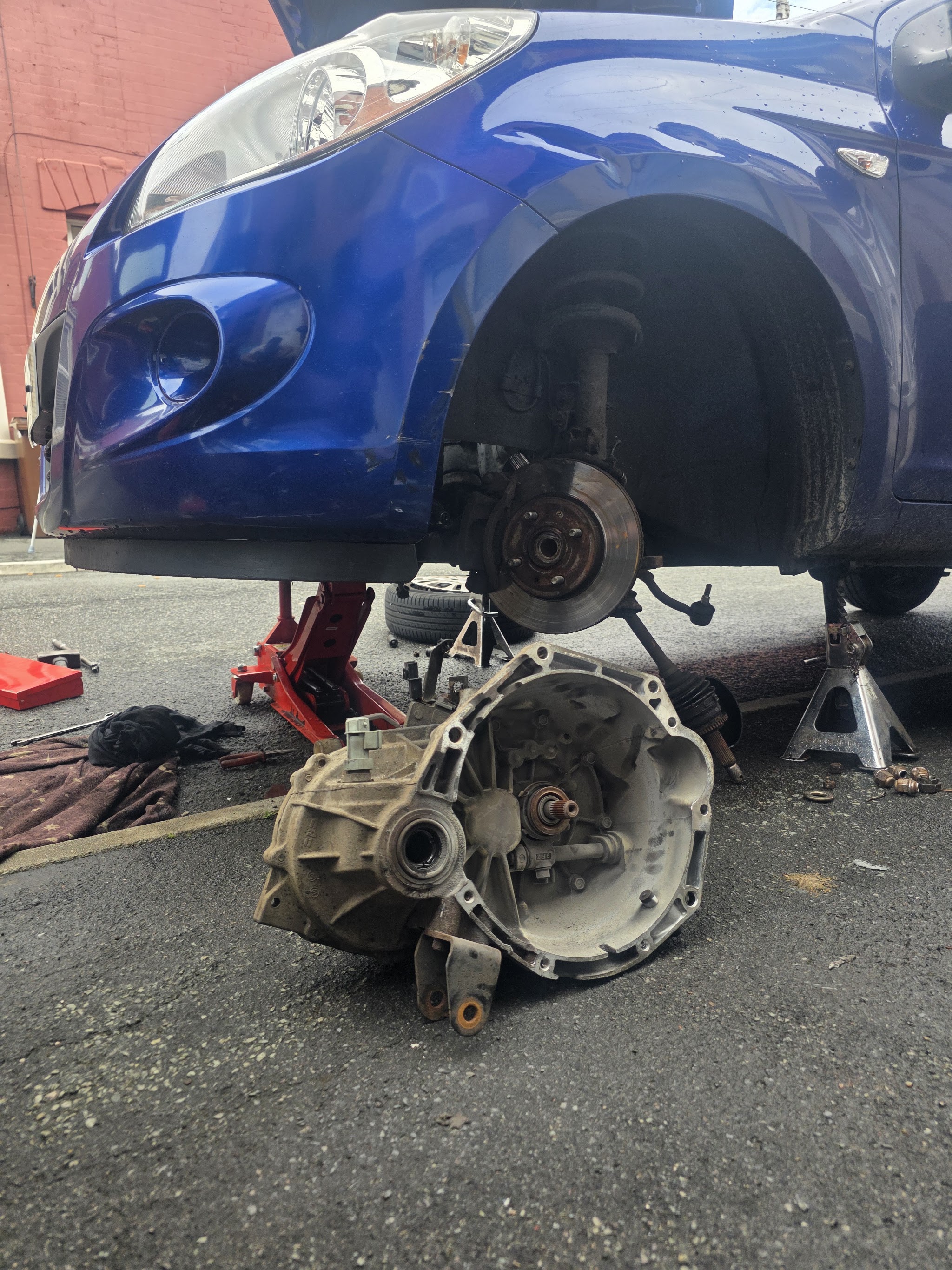 A blue hatchback on a jack and stands at the kerb with its gearbox out on the tarmac beside it
